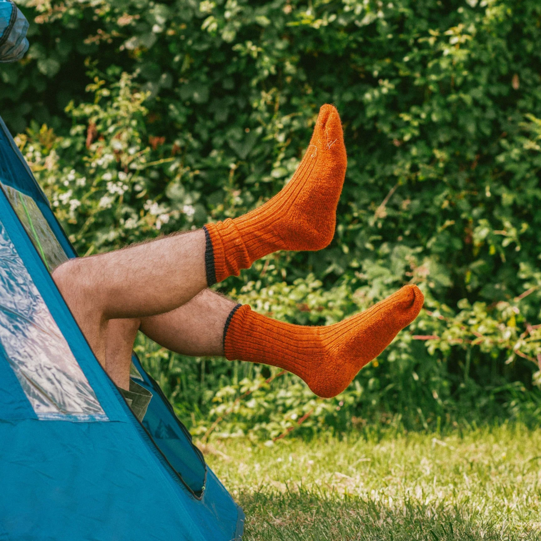 A person sitting in a blue tent outdoors, wearing bright orange ribbed socks against a background of green foliage and grass.