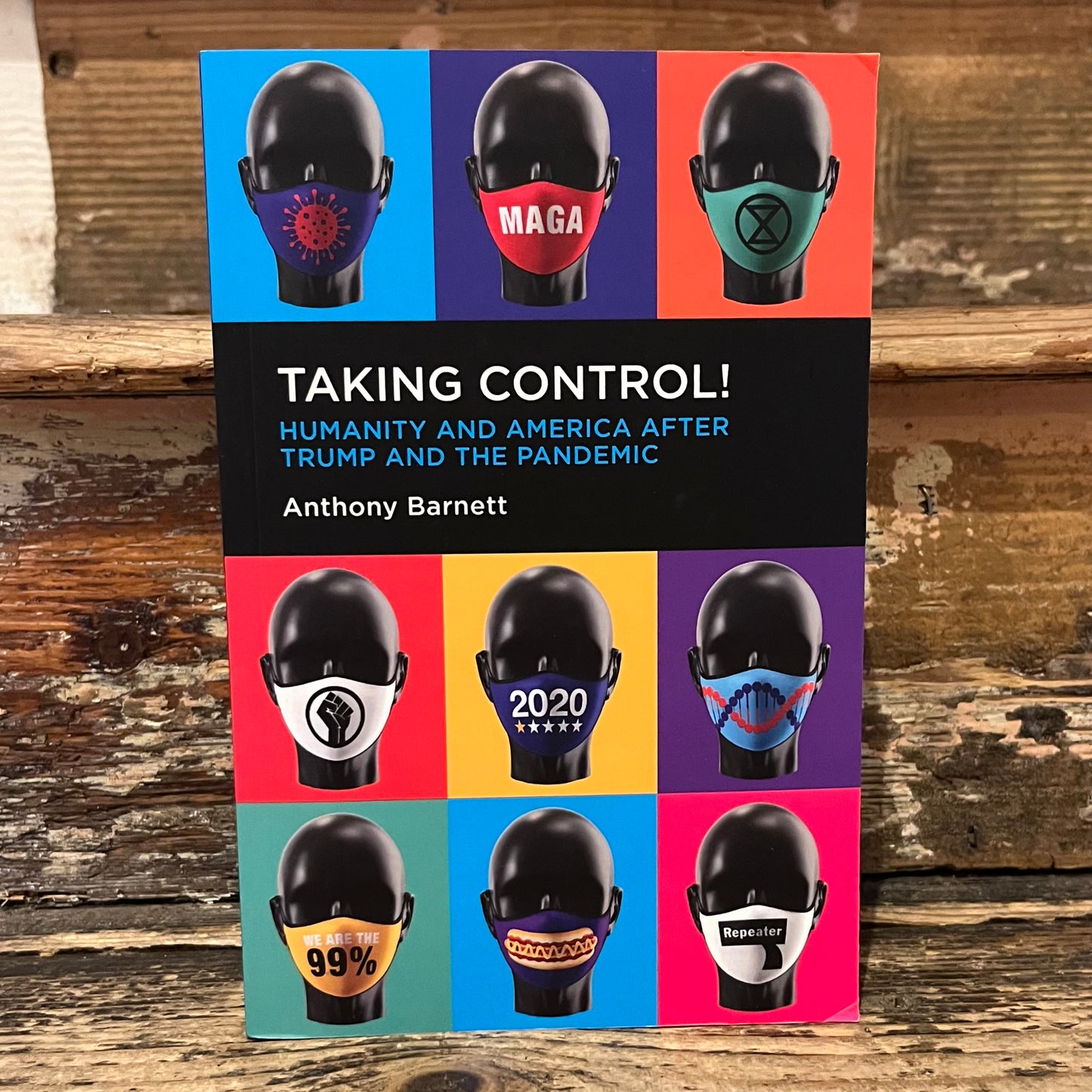 A book titled "Taking Control! Humanity and America After Trump and the Pandemic" by Anthony Barnett, standing upright on a rustic wooden surface. The cover features a grid of nine colorful panels, each showing a black mannequin head wearing a different face mask with political or social slogans.