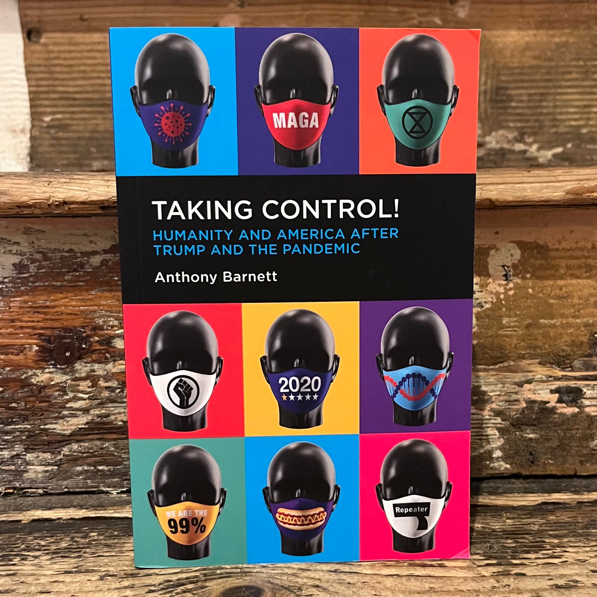 A book titled "Taking Control! Humanity and America After Trump and the Pandemic" by Anthony Barnett, standing upright on a rustic wooden surface. The cover features a grid of nine colorful panels, each showing a black mannequin head wearing a different face mask with political or social slogans.