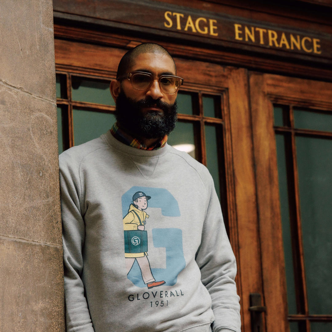 A man with a beard and glasses wearing a light gray crewneck sweatshirt with a graphic of a walking figure and the text 'GLOVERALL 1981', standing in front of a wooden building entrance with 'STAGE ENTRANCE' written above.