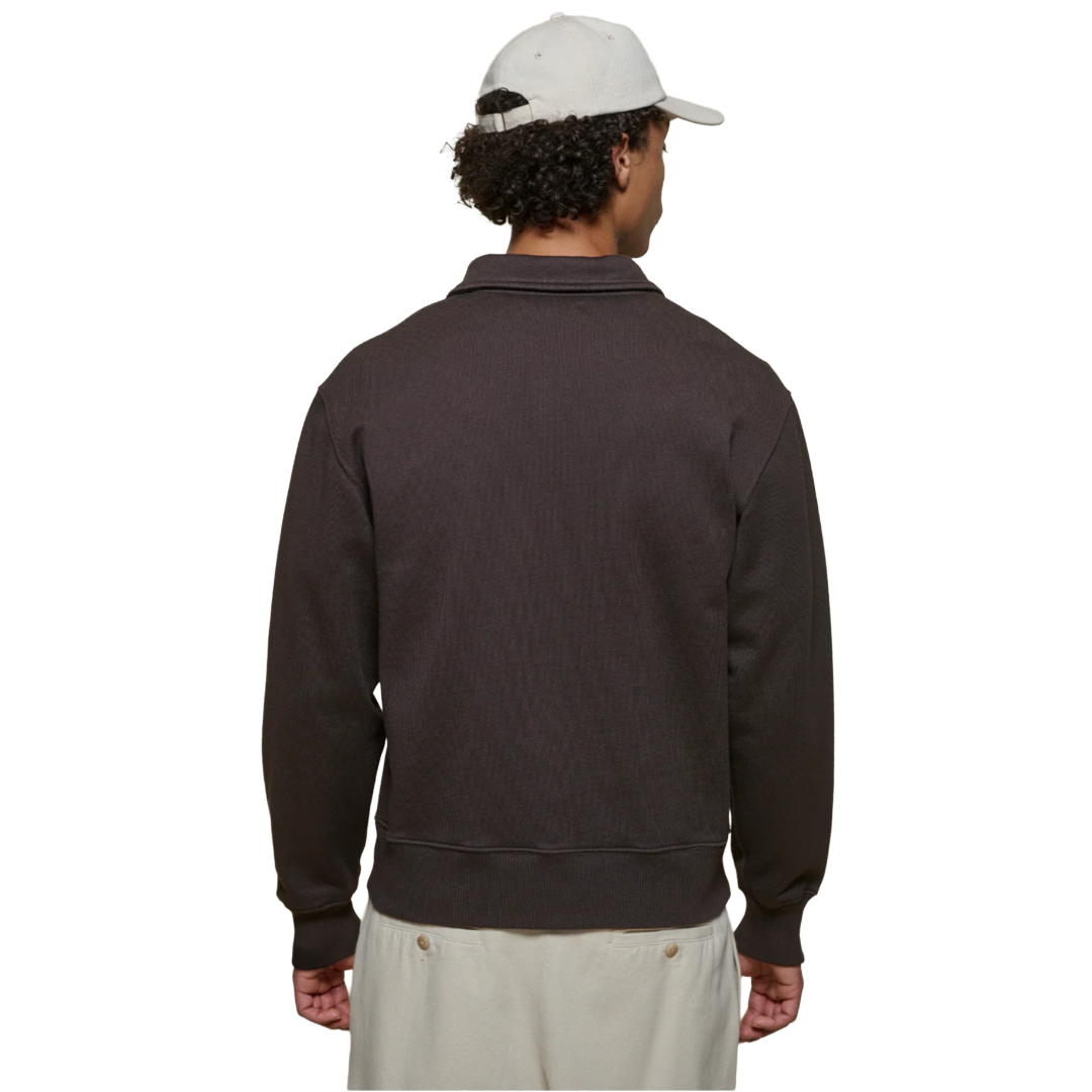 A man viewed from the back wearing a dark brown ribbed knit quarter-zip sweatshirt, a white baseball cap, and off-white pants, set against a plain white background.