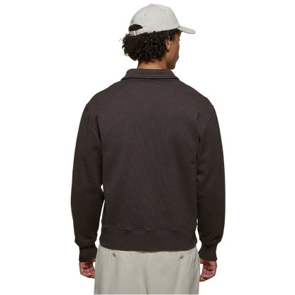 A man viewed from the back wearing a dark brown ribbed knit quarter-zip sweatshirt, a white baseball cap, and off-white pants, set against a plain white background.