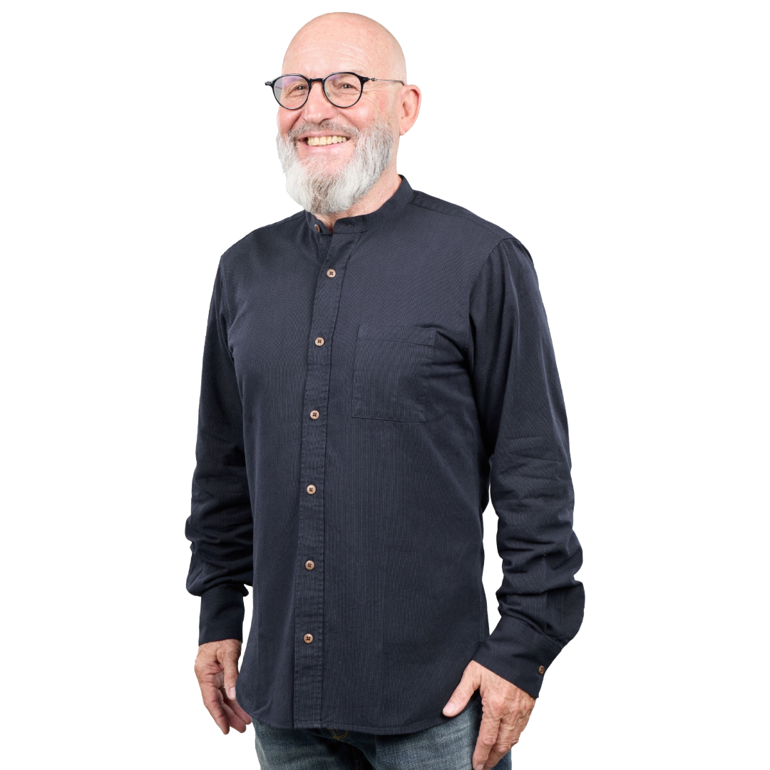 A smiling bald man with a white beard and glasses wearing a dark navy long-sleeve button-up shirt with a mandarin collar, photographed against a solid white background.
