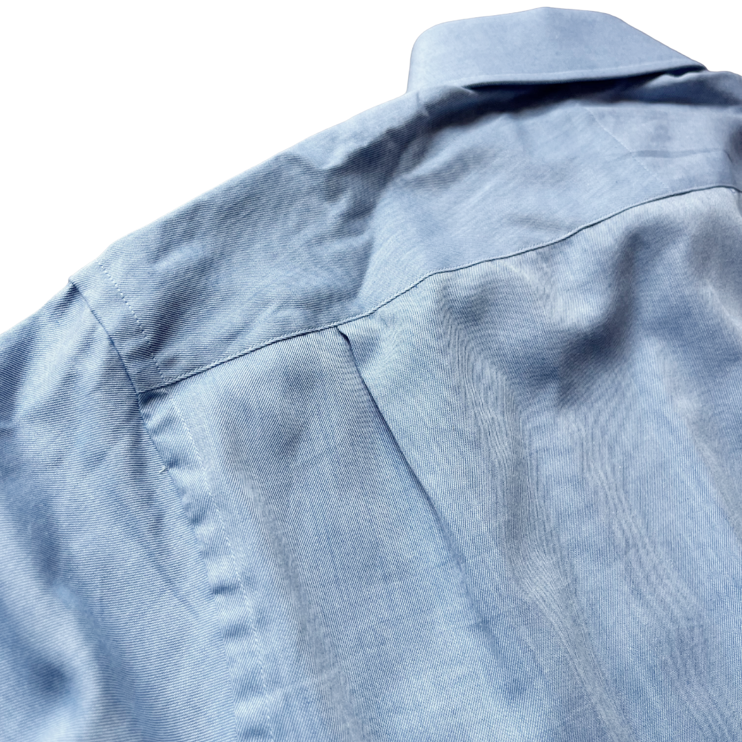 Close-up of a light blue textured fabric garment, showing the collar, shoulder seam, and fine woven texture against a white background.