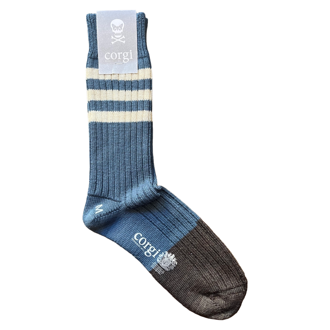 A single crew sock by corgi, featuring a blue ribbed body with two cream stripes near the cuff, a gray cuff label with a skull logo, and a dark charcoal toe and heel.