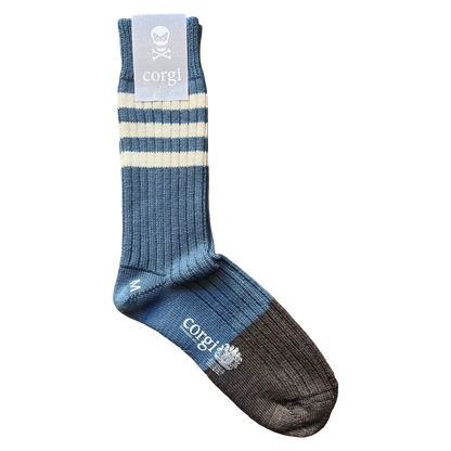 A single crew sock by corgi, featuring a blue ribbed body with two cream stripes near the cuff, a gray cuff label with a skull logo, and a dark charcoal toe and heel.