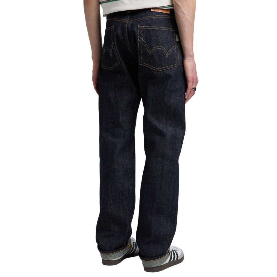 Rear view of a person wearing dark indigo denim jeans with contrast gold stitching, paired with gray and white sneakers, on a solid white background.