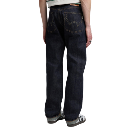 Rear view of a person wearing dark indigo denim jeans with contrast gold stitching, paired with gray and white sneakers, on a solid white background.