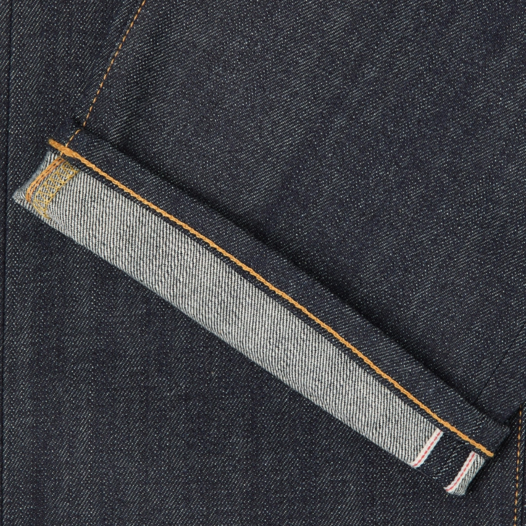 Close-up of a dark indigo denim fabric with a folded hem showing a lighter gray inner lining and orange contrast stitching.