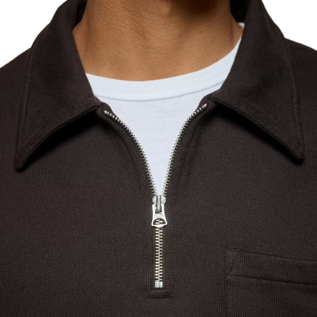 Close-up of a person wearing a dark brown ribbed knit quarter-zip pullover with a collar, layered over a white t-shirt. The zipper pull features a small silver-toned metal logo.