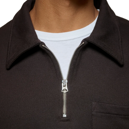 Close-up of a person wearing a dark brown ribbed knit quarter-zip pullover with a collar, layered over a white t-shirt. The zipper pull features a small silver-toned metal logo.