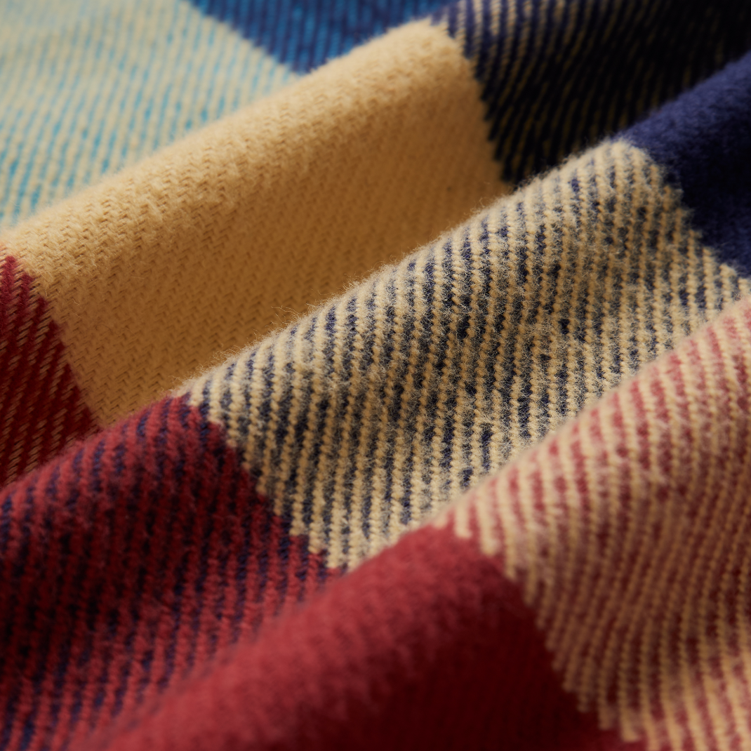 Close-up of a soft woven plaid fabric with large check blocks in cream, navy, blue, burgundy red, and muted pink, showing the textile texture and weave.