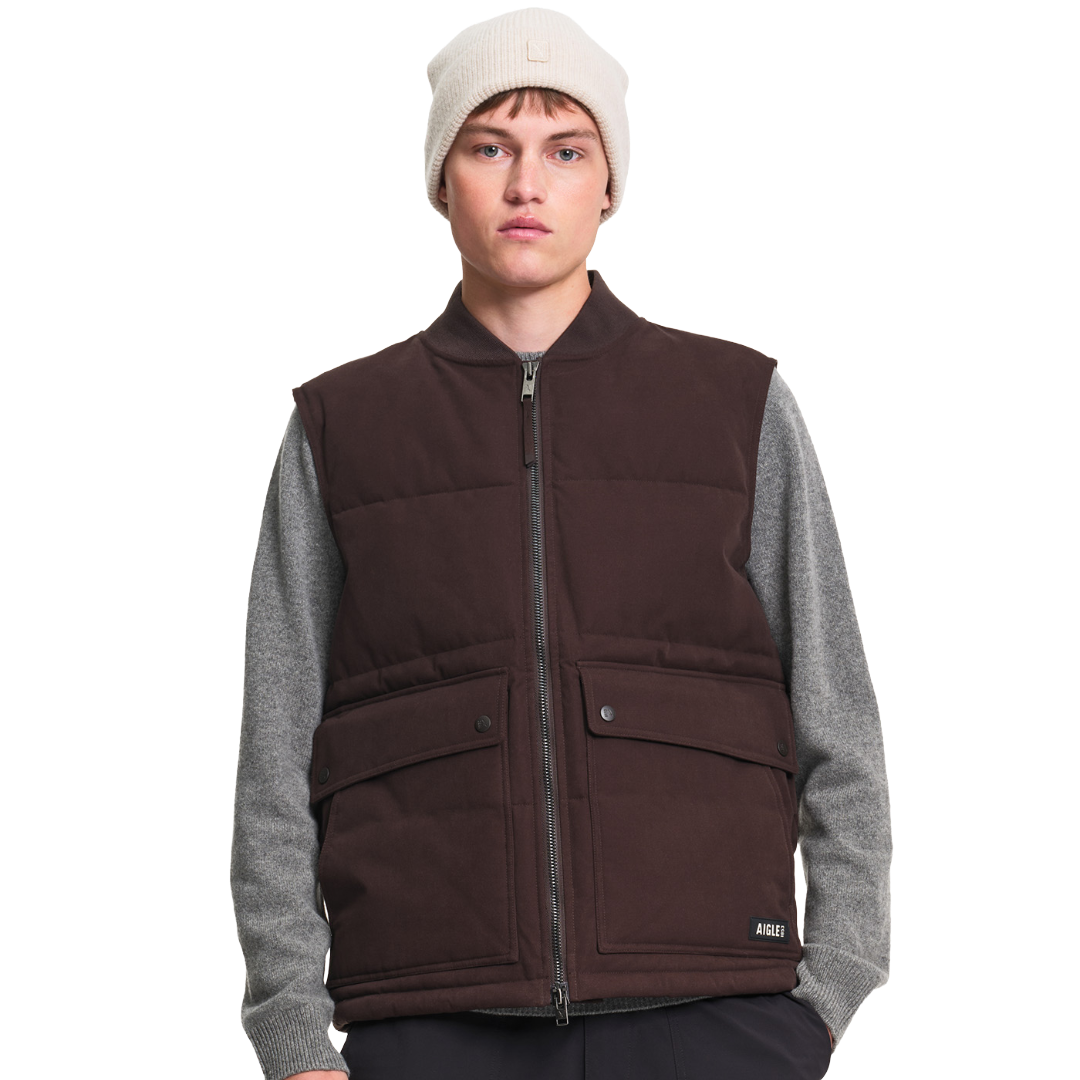 A male model wearing a dark brown puffer vest over a gray long-sleeve shirt, paired with a cream beanie, photographed against a solid white background.