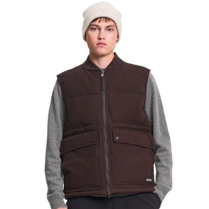 A male model wearing a dark brown puffer vest over a gray long-sleeve shirt, paired with a cream beanie, photographed against a solid white background.