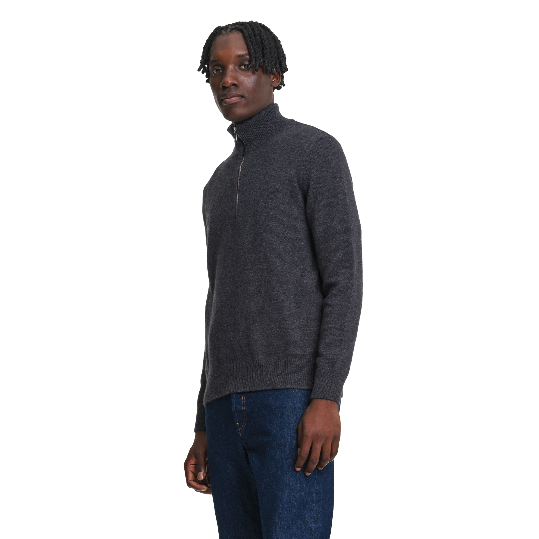 A man wearing a dark gray quarter-zip knit sweater and blue jeans, standing against a plain white background.