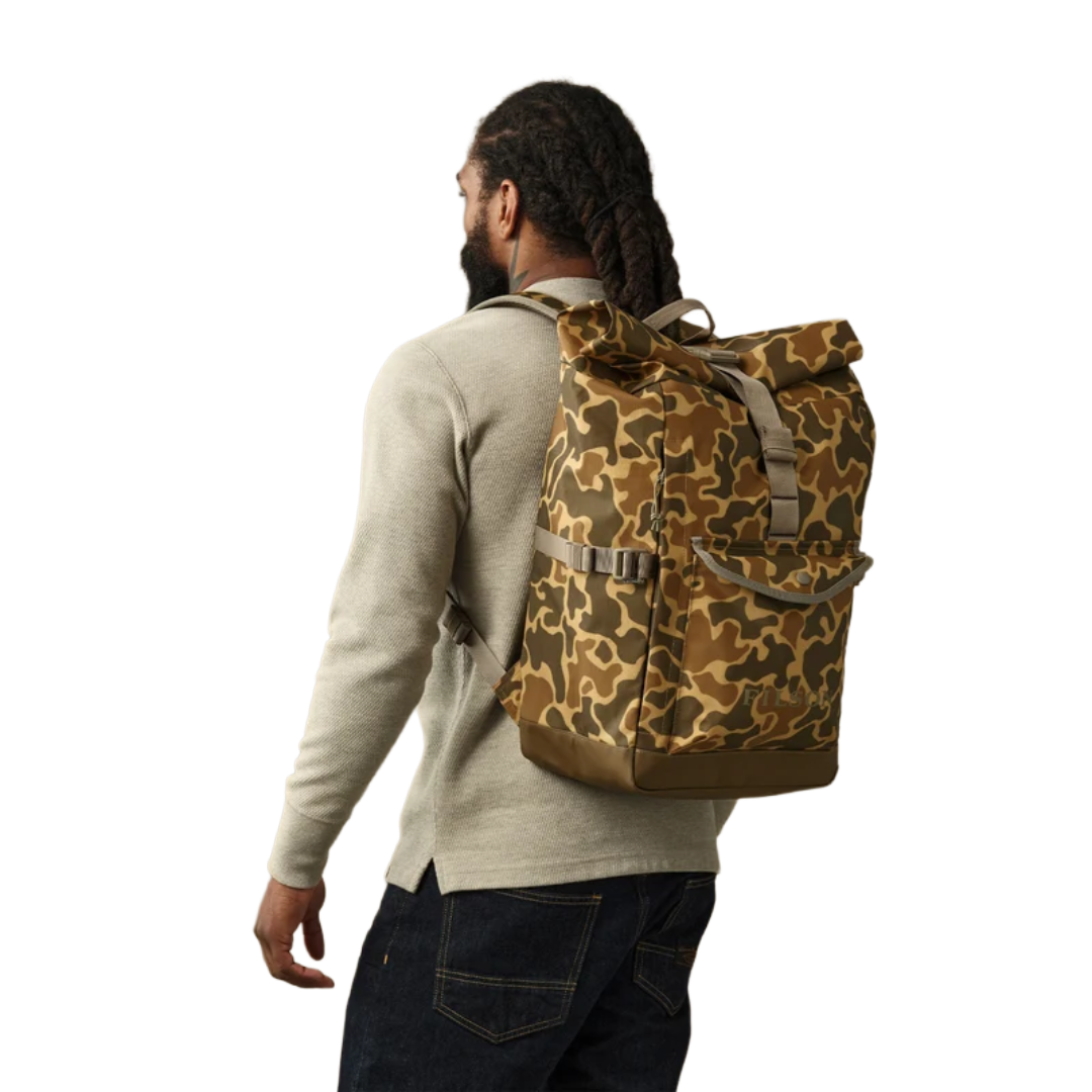 A man wearing a beige sweater and dark jeans is shown from behind, carrying a brown and tan camouflage backpack on his back against a plain white background.