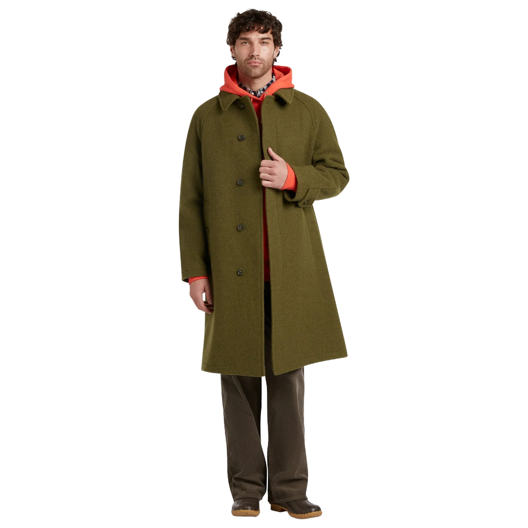 A man modeling a long olive green wool-blend coat over a bright orange hoodie, brown pants, and brown shoes, photographed against a solid white background.