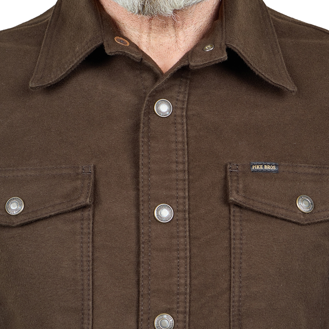 Close-up of a brown waxed canvas button-up shirt by Jicks Bros., featuring a collar, silver snap buttons, and chest pockets.