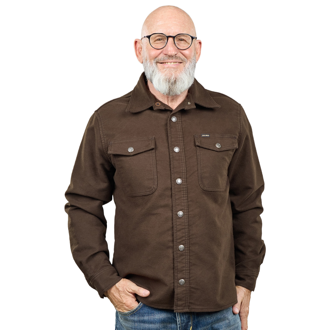 A smiling man with glasses, a gray beard, and a bald head wearing a brown button-up shirt with two chest pockets, paired with blue jeans, against a plain white background.