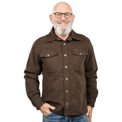 A smiling man with glasses, a gray beard, and a bald head wearing a brown button-up shirt with two chest pockets, paired with blue jeans, against a plain white background.