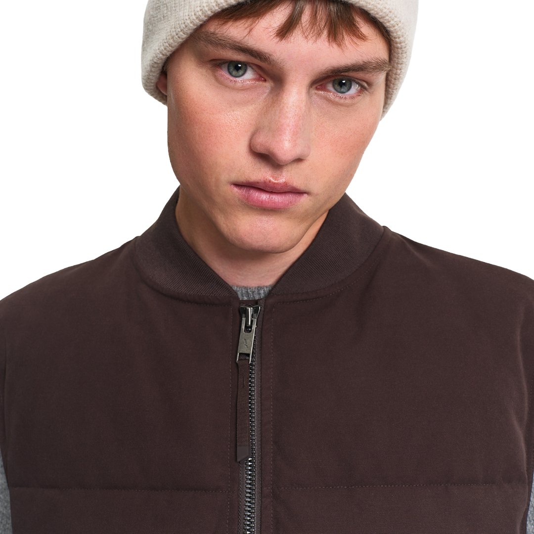 Close-up of a person wearing a dark brown zip-front vest over a gray shirt, with a light beige knit beanie, against a plain white background.