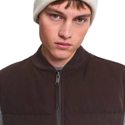 Close-up of a person wearing a dark brown zip-front vest over a gray shirt, with a light beige knit beanie, against a plain white background.
