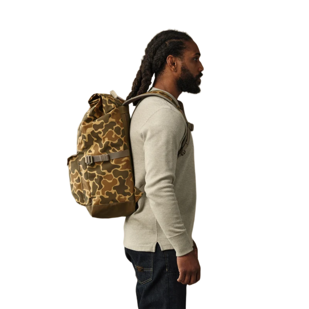 A man in a beige sweater and dark jeans shown from the side, wearing a brown and tan camouflage backpack against a plain white background.
