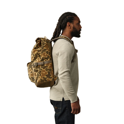 A man in a beige sweater and dark jeans shown from the side, wearing a brown and tan camouflage backpack against a plain white background.