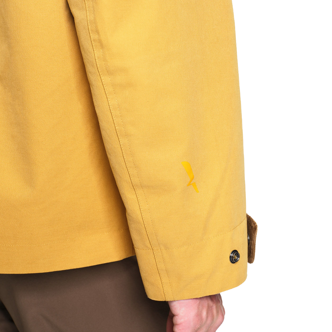 Close-up of a mustard yellow jacket sleeve with a small yellow embroidered logo, showing a hand adjusting a brown button cuff against a white background.