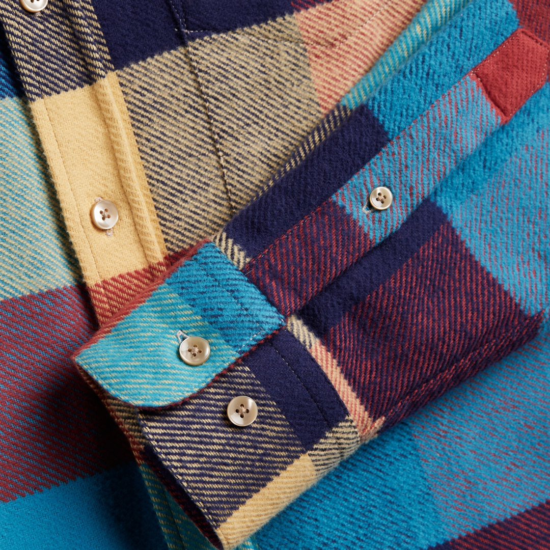 Close-up of a plaid flannel shirt fabric with visible buttons and a folded cuff, showing multiple colors including blue, navy, red, beige, and yellow in a tartan pattern.