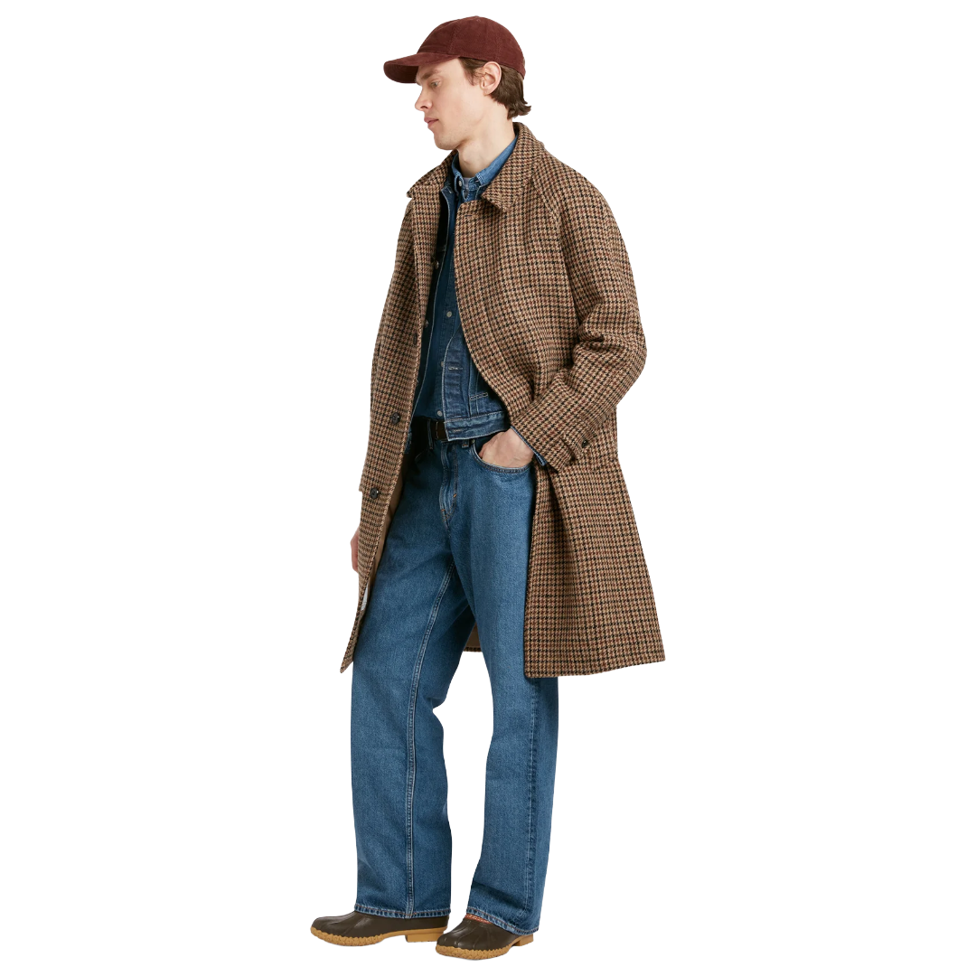 A model wearing a brown and beige plaid overcoat, blue denim shirt and jeans, a maroon baseball cap, and brown shoes, shown in profile against a solid white background.