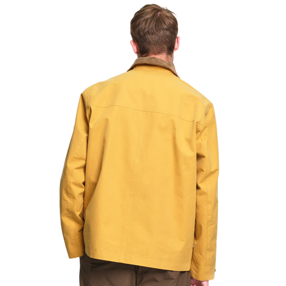 A man viewed from the back wearing a mustard yellow utility jacket with a brown collar, set against a plain white background.