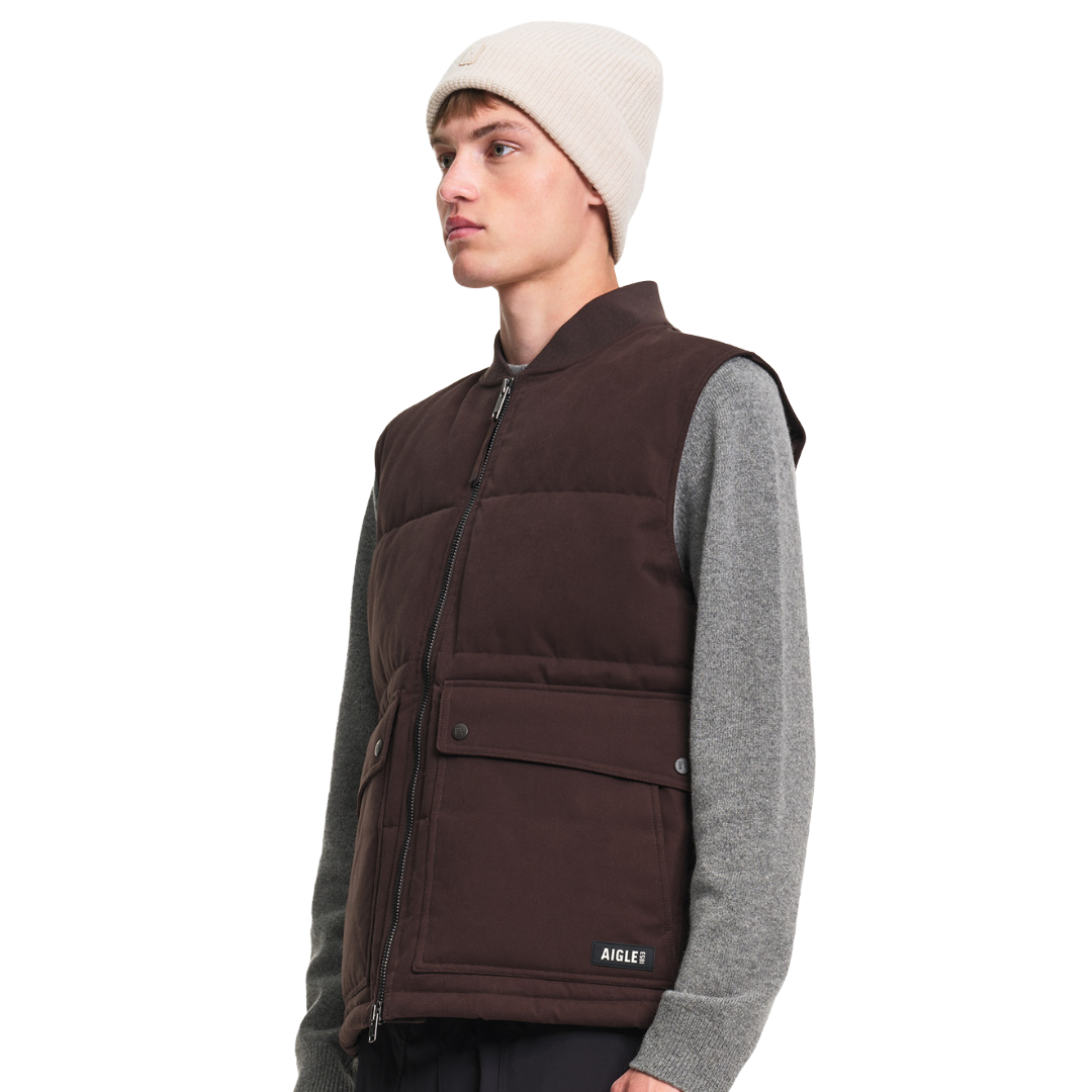 A model wearing a dark brown puffer vest over a gray long-sleeve shirt and a cream beanie, shown from a side profile against a solid white background.