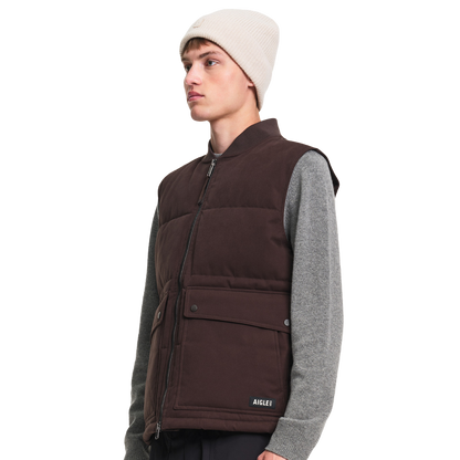 A model wearing a dark brown puffer vest over a gray long-sleeve shirt and a cream beanie, shown from a side profile against a solid white background.