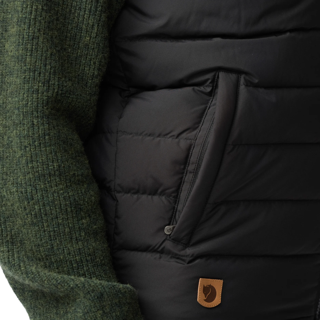 Close-up of a black puffer vest with horizontal quilting, a side zip pocket, and a small brown Fjäll logo patch, worn over a dark green ribbed knit sweater.