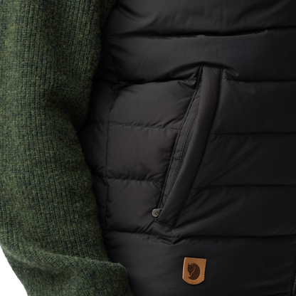 Close-up of a black puffer vest with horizontal quilting, a side zip pocket, and a small brown Fjäll logo patch, worn over a dark green ribbed knit sweater.