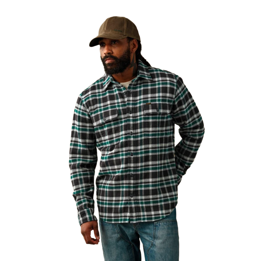 A man wearing a long-sleeve button-up plaid flannel shirt in black, green, and white, paired with blue jeans and a brown baseball cap, set against a plain white background.