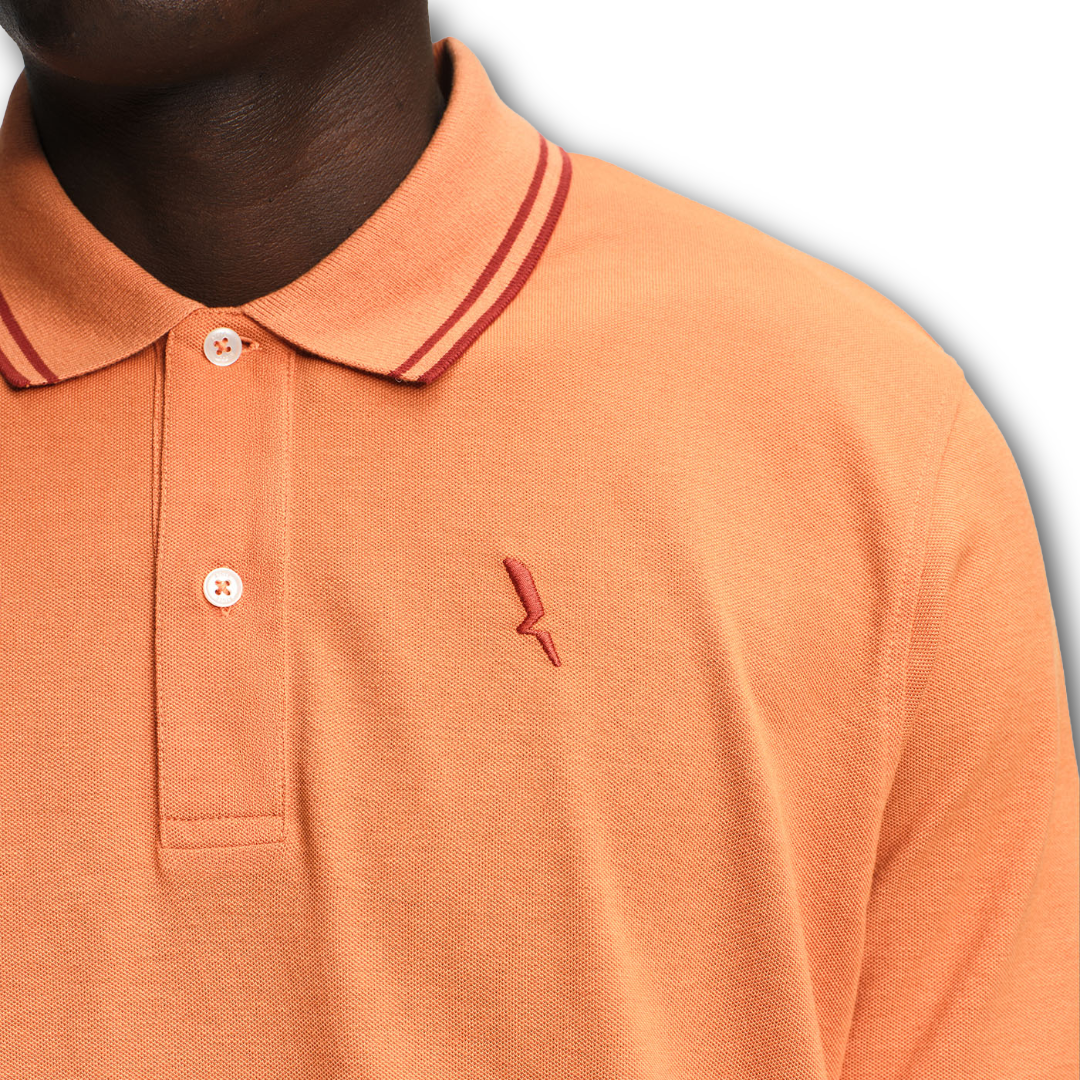Close-up of an Aigle orange polo shirt with a small logo on a gray background sold at Regent in Salisbury