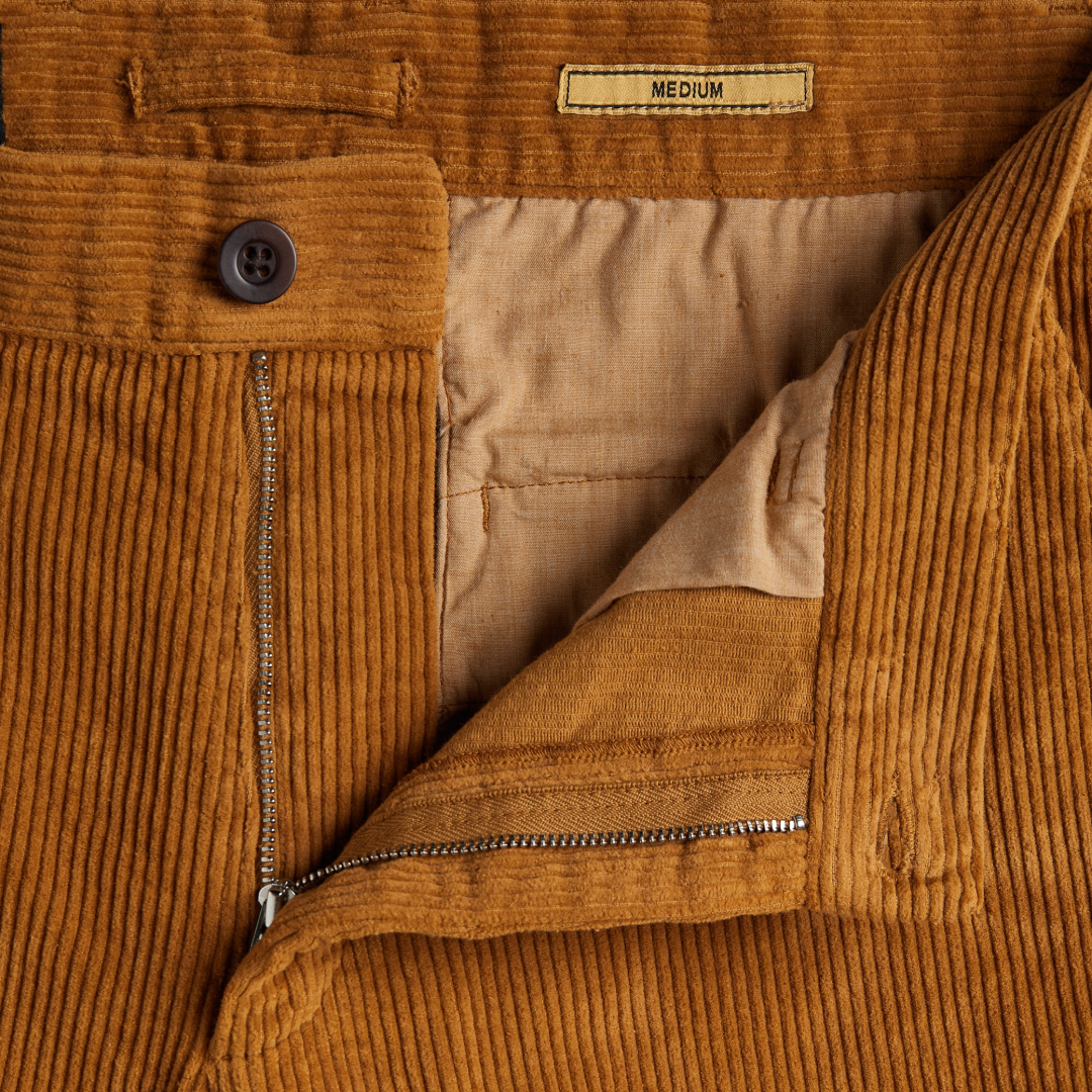 Close-up of a mustard corduroy jacket with a brown button, silver zipper, and tan interior lining. A label at the waistband reads 'MEDIUM'.