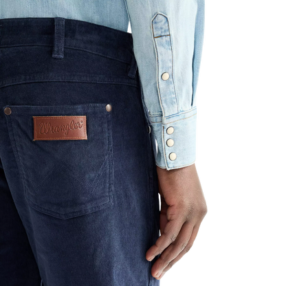 Close-up of a person wearing dark blue corduroy jeans with a brown leather Wrangler patch on the back pocket, paired with a light blue denim shirt against a white background.