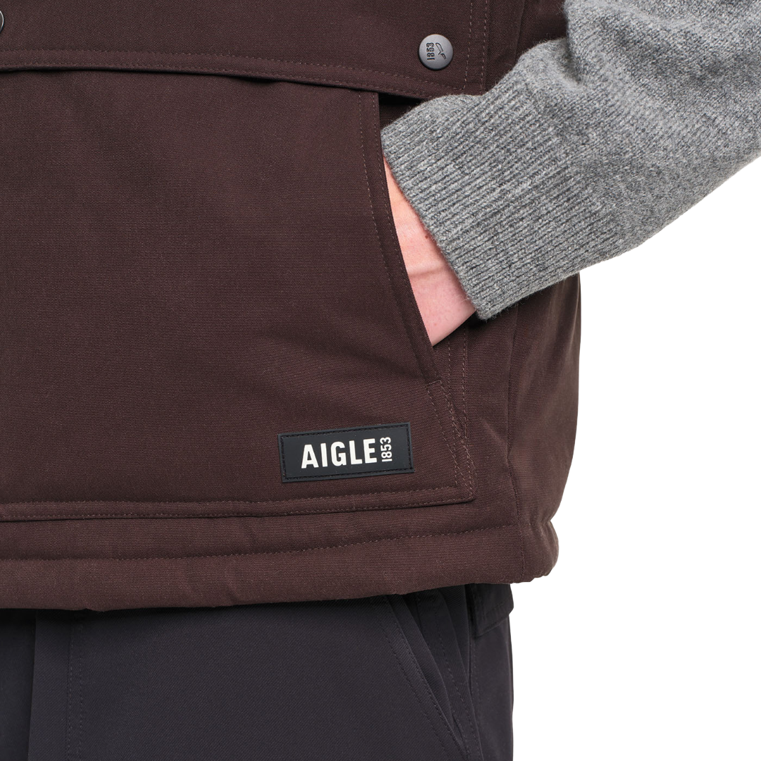 Close-up of a person wearing a dark brown Aigle 1853 jacket, showing a hand in a pocket and a gray knit sleeve, on a white background.