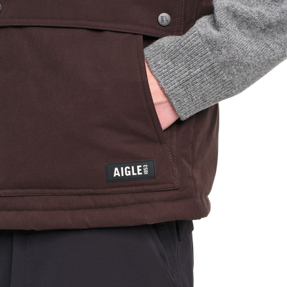 Close-up of a person wearing a dark brown Aigle 1853 jacket, showing a hand in a pocket and a gray knit sleeve, on a white background.