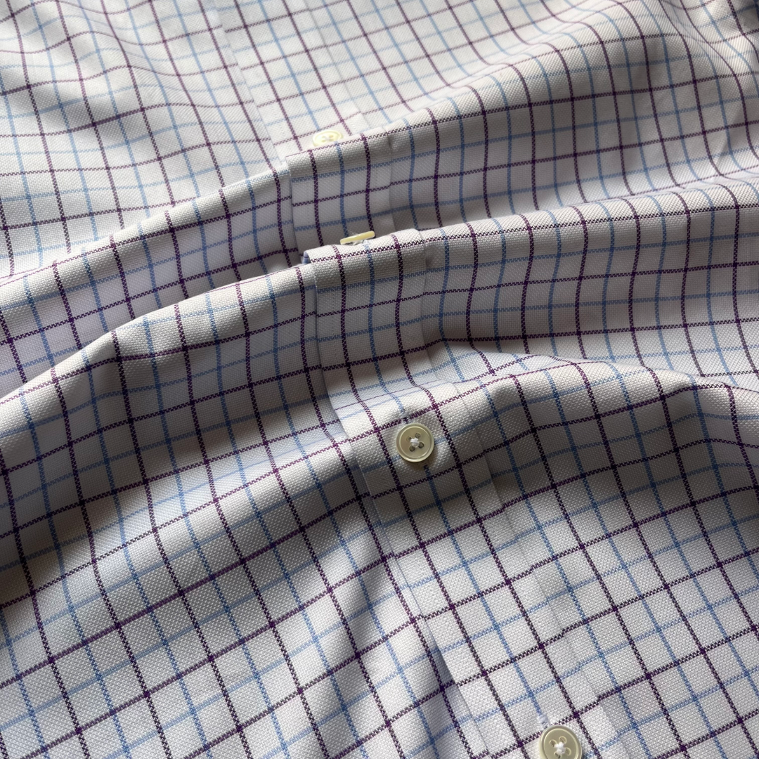 A close-up of a white button-up shirt fabric with a fine windowpane plaid pattern in purple and light blue, featuring cream-colored buttons.