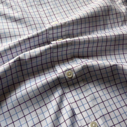 A close-up of a white button-up shirt fabric with a fine windowpane plaid pattern in purple and light blue, featuring cream-colored buttons.