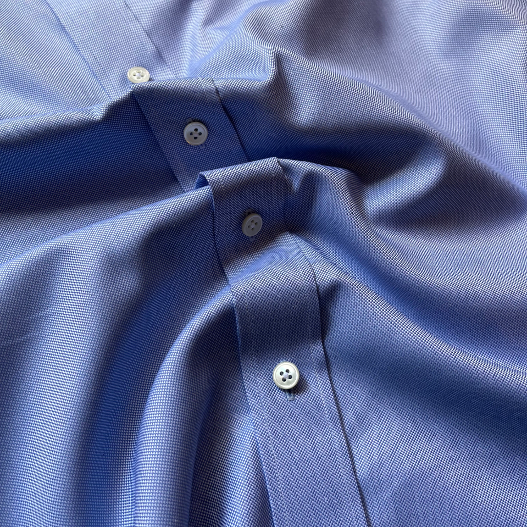 Close-up of a blue micro-patterned button-down shirt with white buttons, showing the fine fabric texture and collar details.