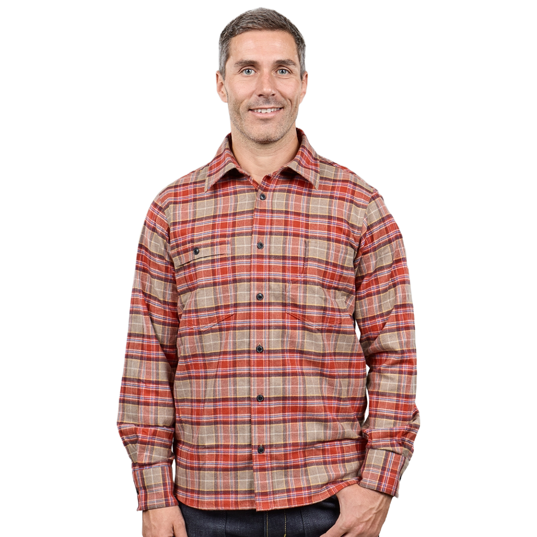 A man wearing a long-sleeve button-up plaid shirt in red, orange, and beige tones, paired with dark jeans, photographed against a solid white background.