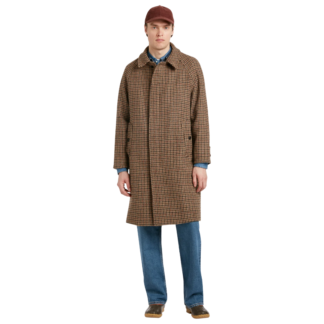 A man modeling a brown and black checkered mid-length overcoat, paired with blue jeans, a blue patterned shirt, and a brown cap, against a solid white background.