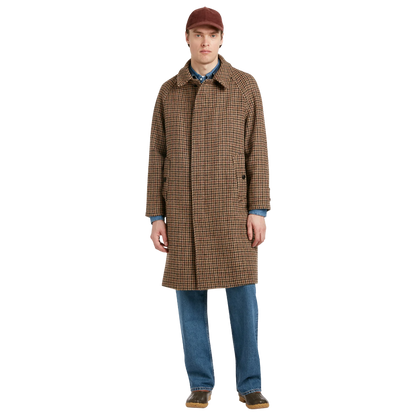 A man modeling a brown and black checkered mid-length overcoat, paired with blue jeans, a blue patterned shirt, and a brown cap, against a solid white background.