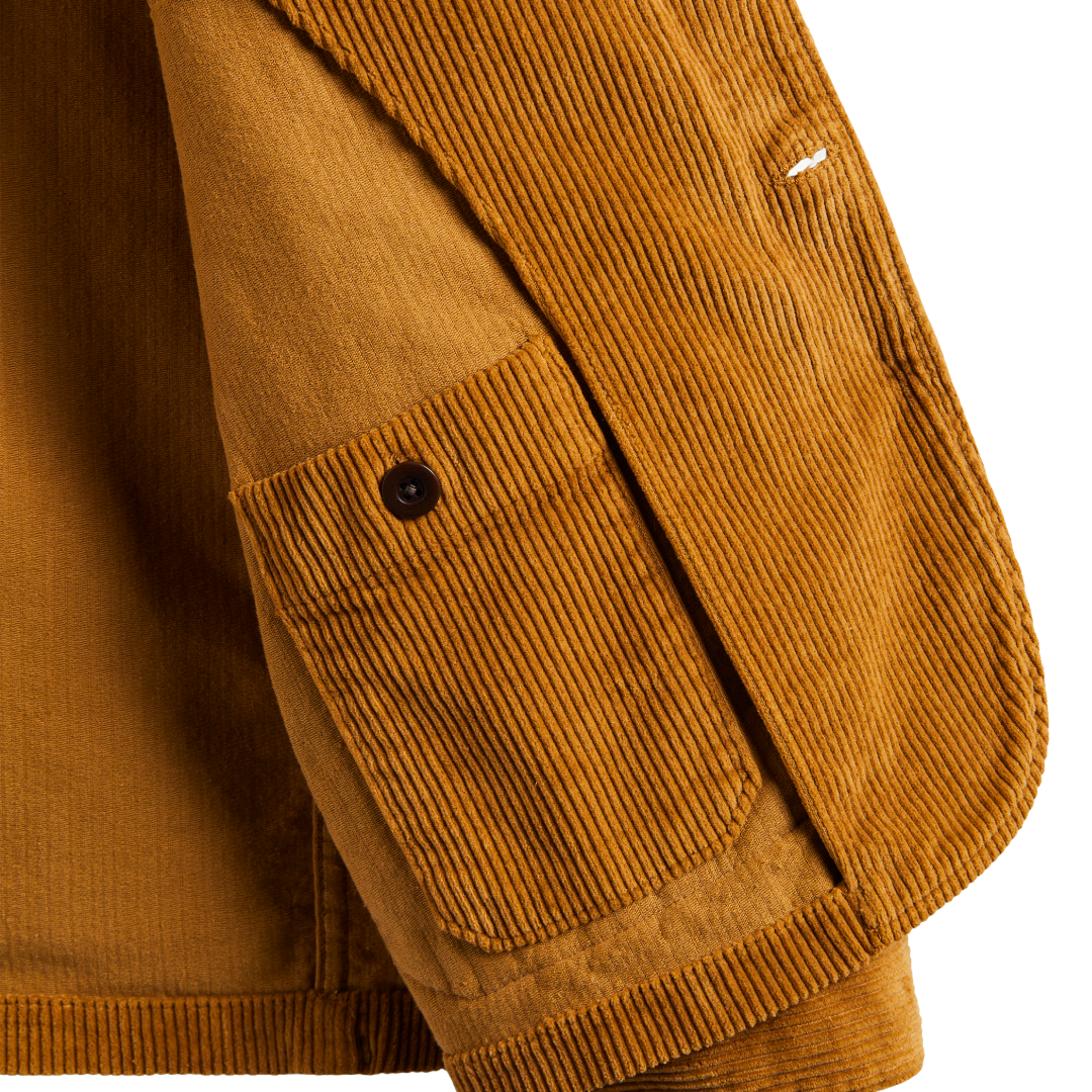 Close-up detail of a mustard brown corduroy jacket showing a buttoned chest pocket and ribbed fabric texture against a white background.