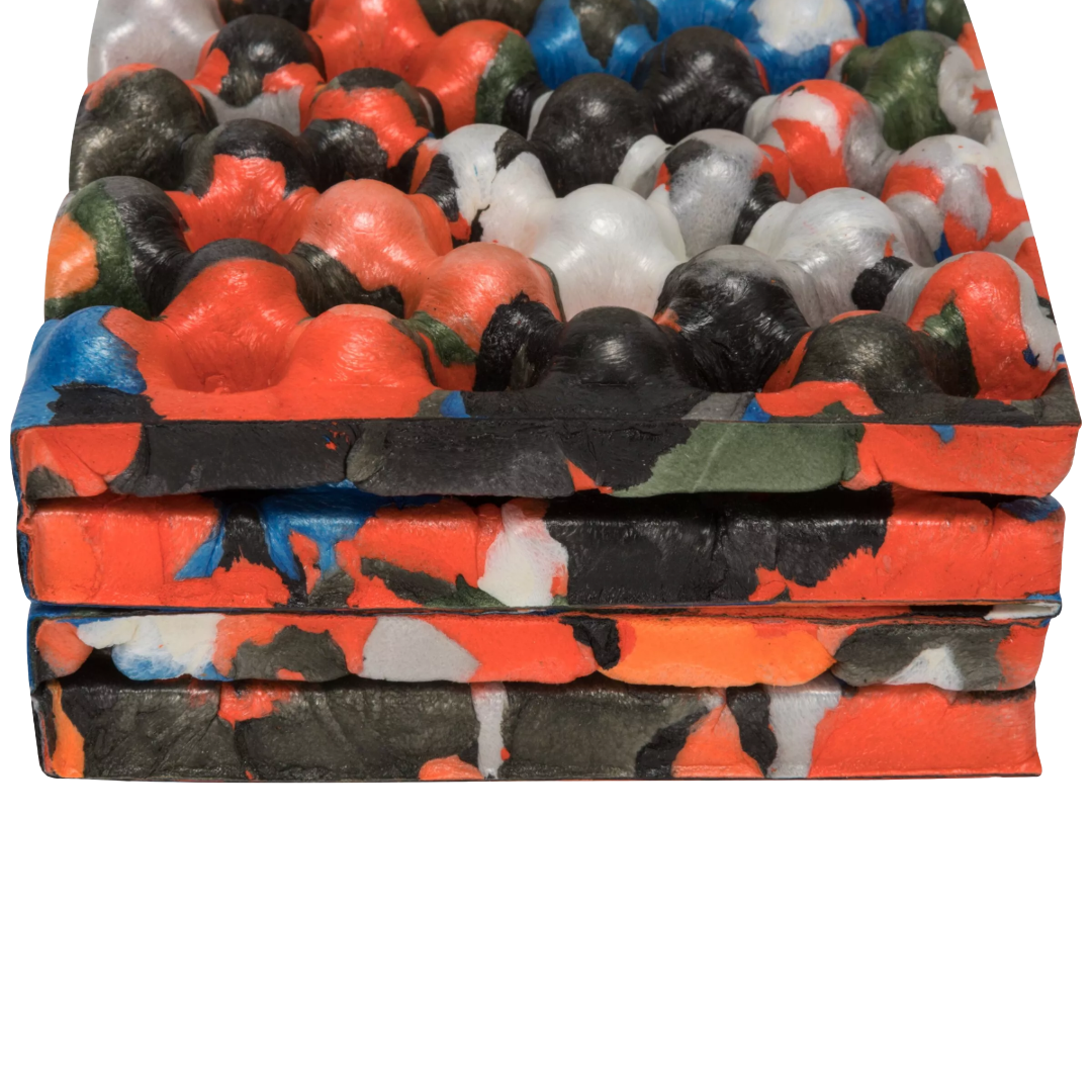 A close-up of a stack of thick, textured foam mats with a bold abstract paint-splatter pattern in orange, black, red, blue, green, and white, set against a plain white background.